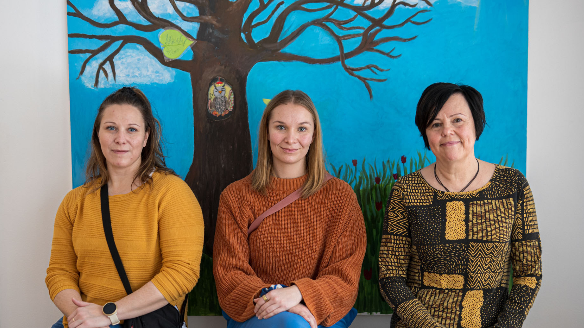 Mervi Taipalus, Roosa-Maria Routi ja Taina Väinölä istuvat kotiutumispuun edessä, johon kerätään tsemppiviestejä sijaishuollosta kotiutuvilta nuorilta saapuville nuorille.